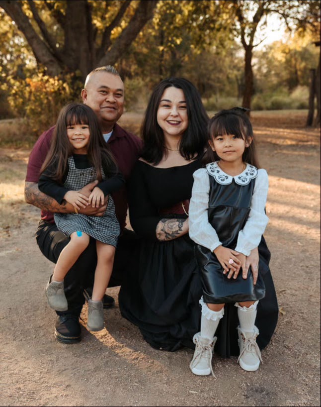 "Nicole Phommanorat with her husband and two young daughters, smiling outdoors in a park setting with golden fall light and trees in the background."