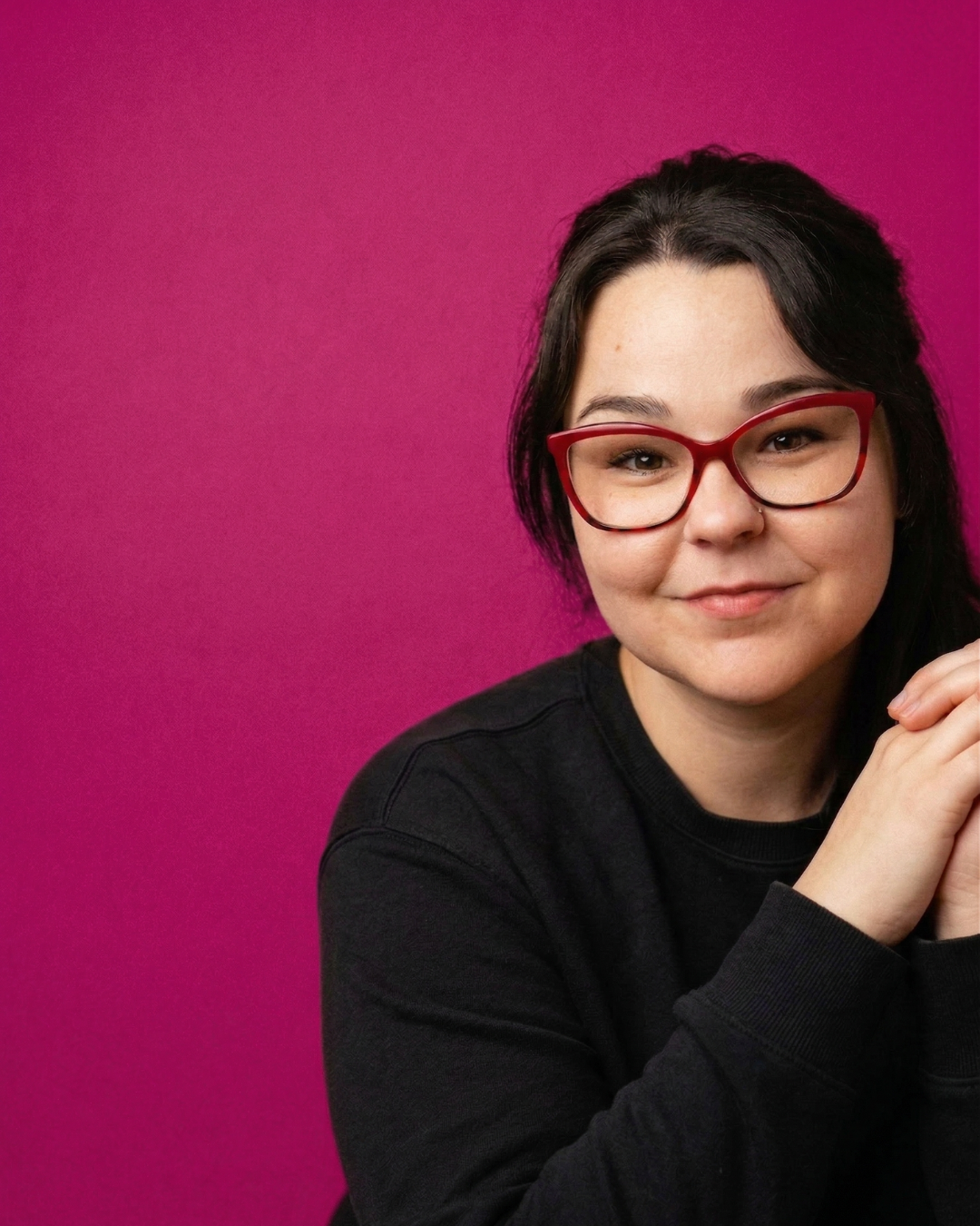 Nicole Phommanorat, founder of We Thrive Collective, wearing red cat-eye glasses and a black sweater against a magenta background