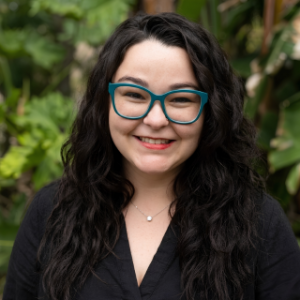 Nicole Phommanorat, founder of We Thrive Collective, wearing red cat-eye glasses and a black sweater against a green tropical background
